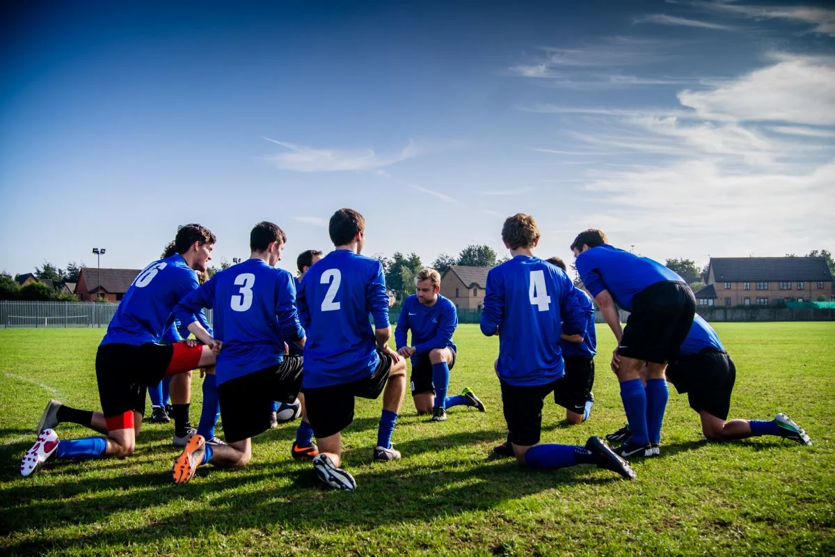 A football team in blue kits having a discussion on a grassy pitch with houses in the background on a clear day.